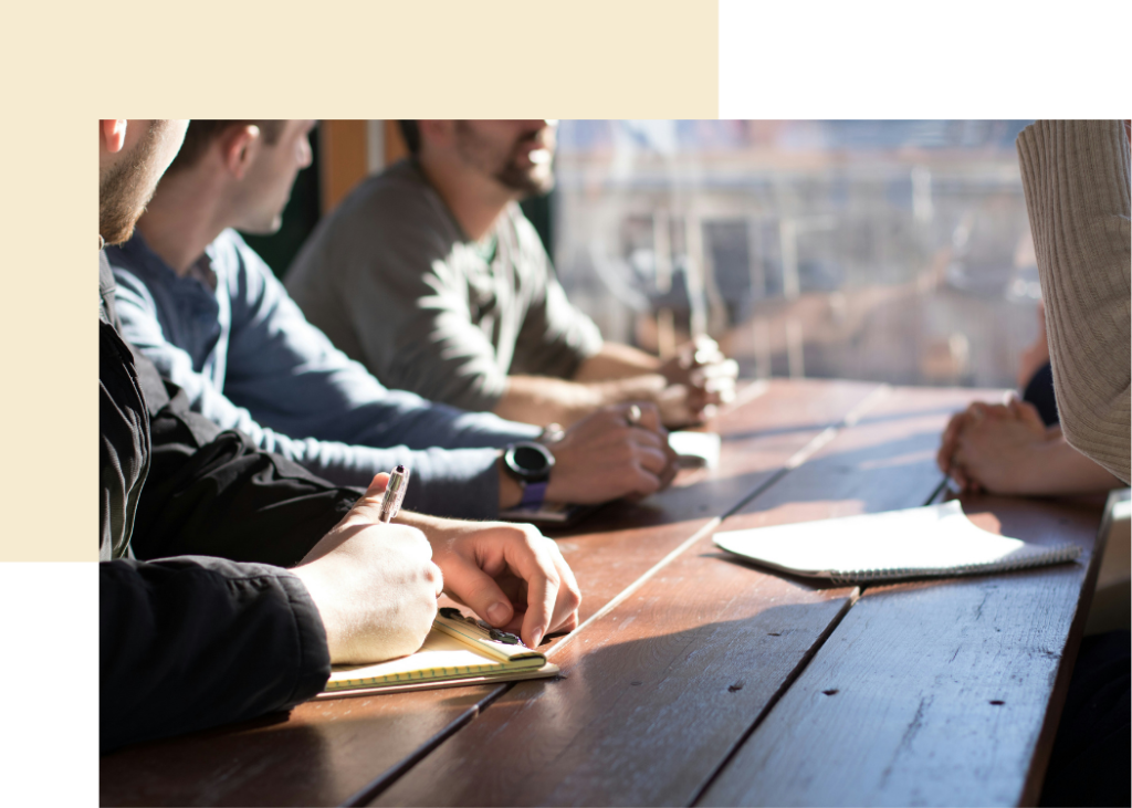 Several people sitting around a wooden table with notepads. You cannot see their faces, but you can see a few mens chins and other peoples arms and hands.
