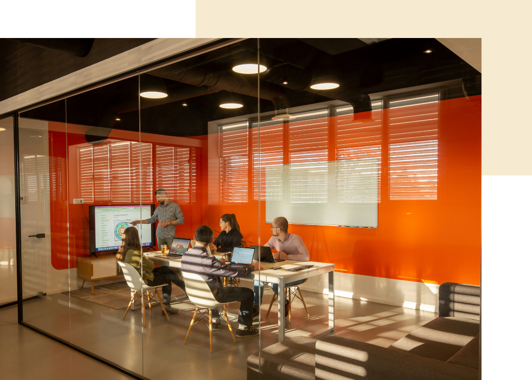 A red conference room with a glass wall. Inside the room five people are working. One is standing at a TV and the other four are sitting with laptops open. 
