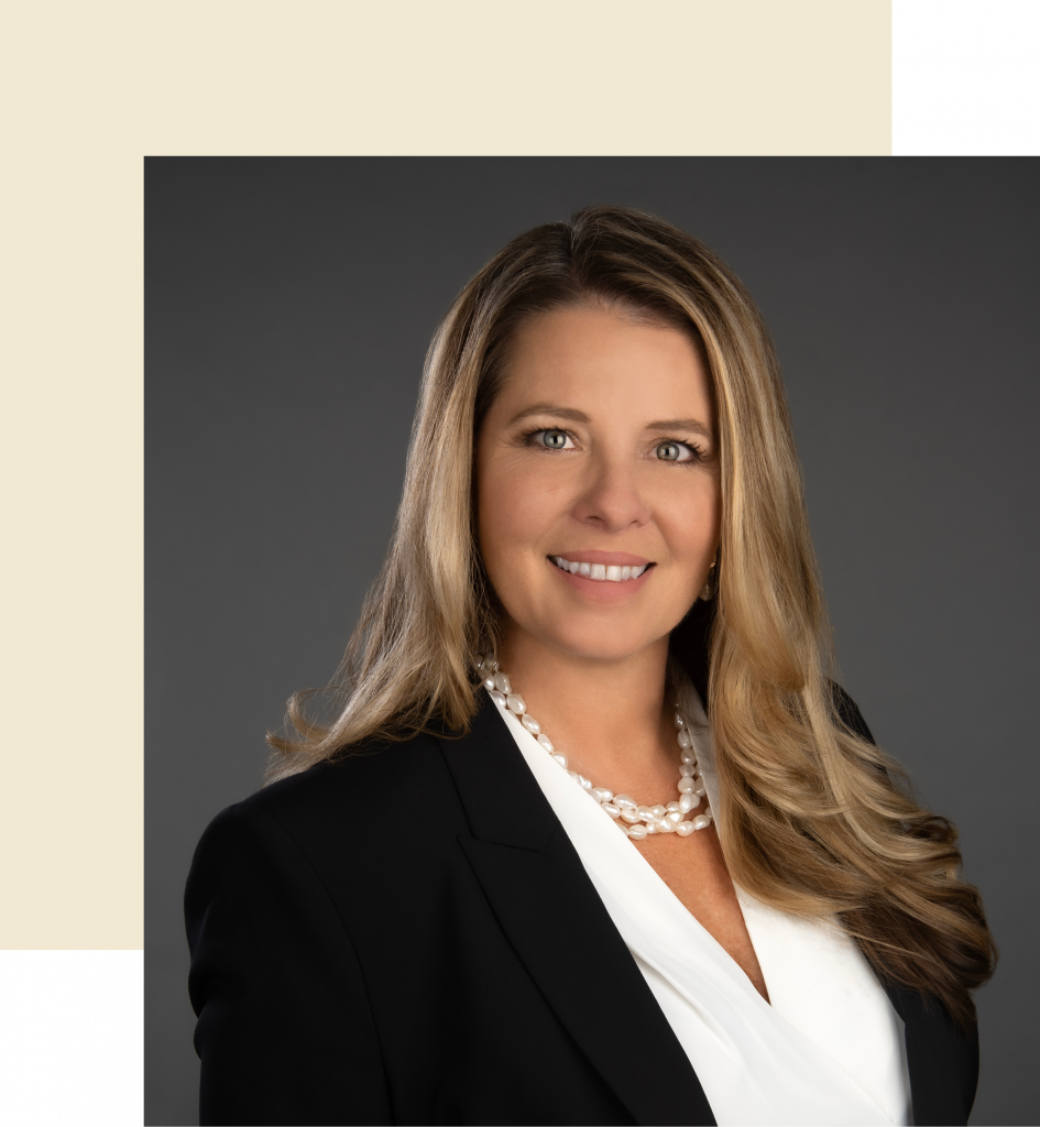 Michelle Baker, a white woman with long blonde hair wearing a pearl necklace, a white blouse, and a black suit jacket in front of a dark gray background.