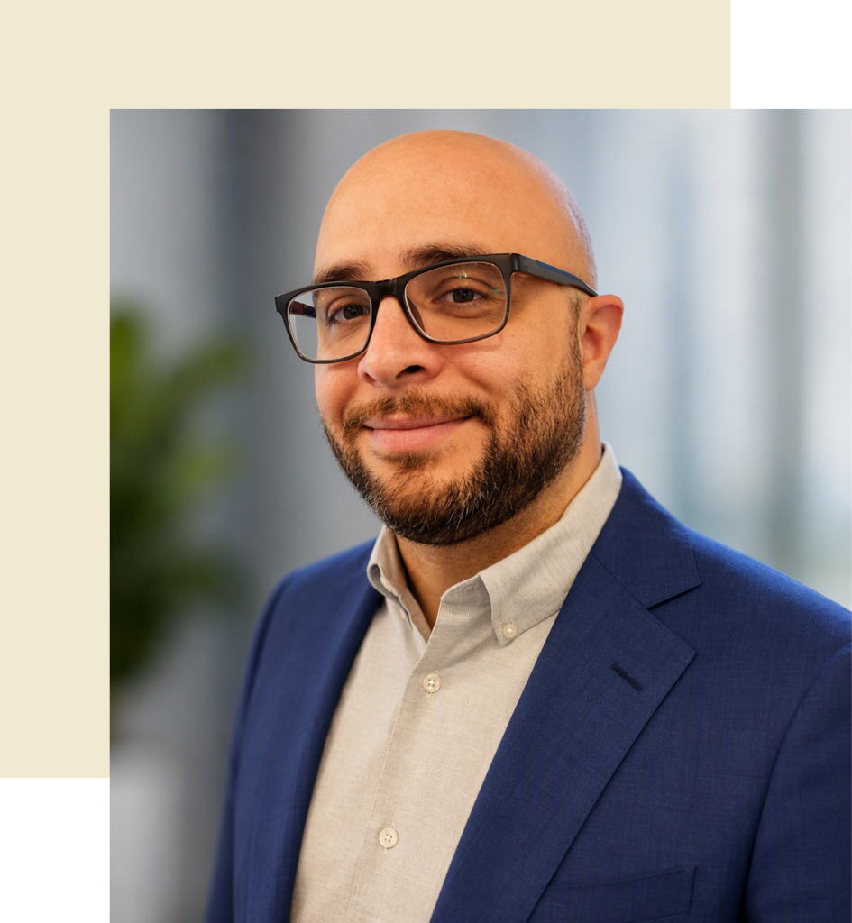 Josh Lecure, a bald white man with glasses and a beard wearing a light gray button down and a navy blue suit jacket in front of a blurred office background.